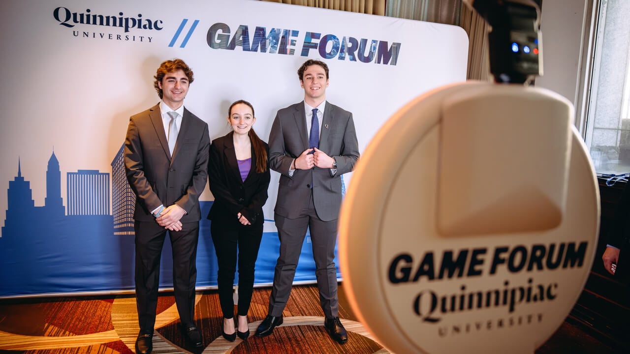 Three students in suits pose for a photo in front of a GAME Forum backdrop