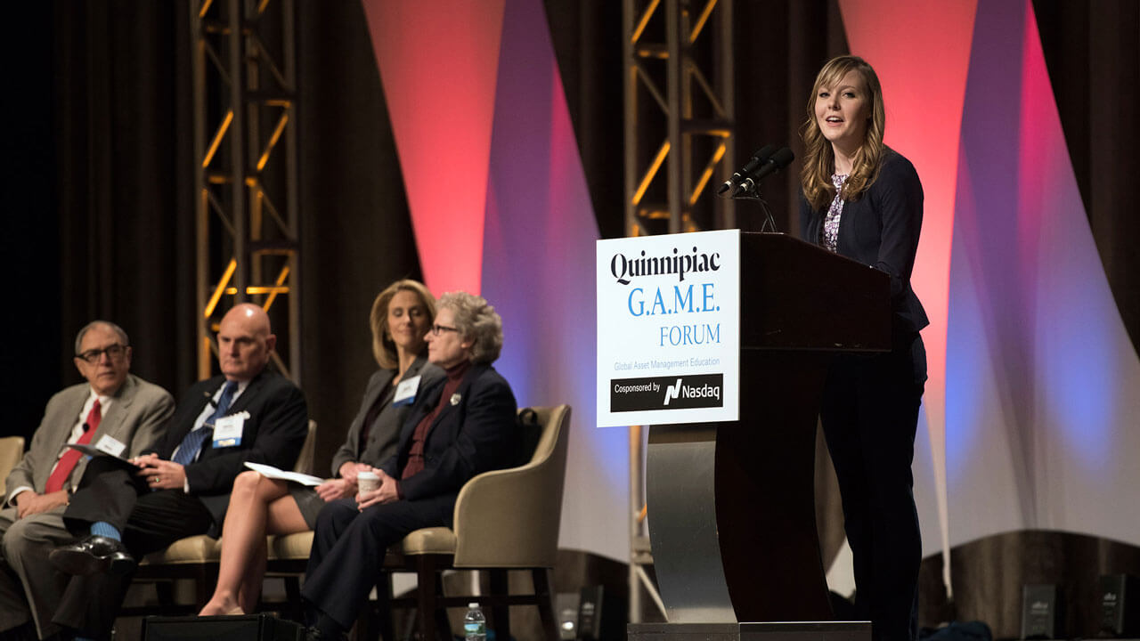A student speaks at a podium on a colorful stage during GAME Forum