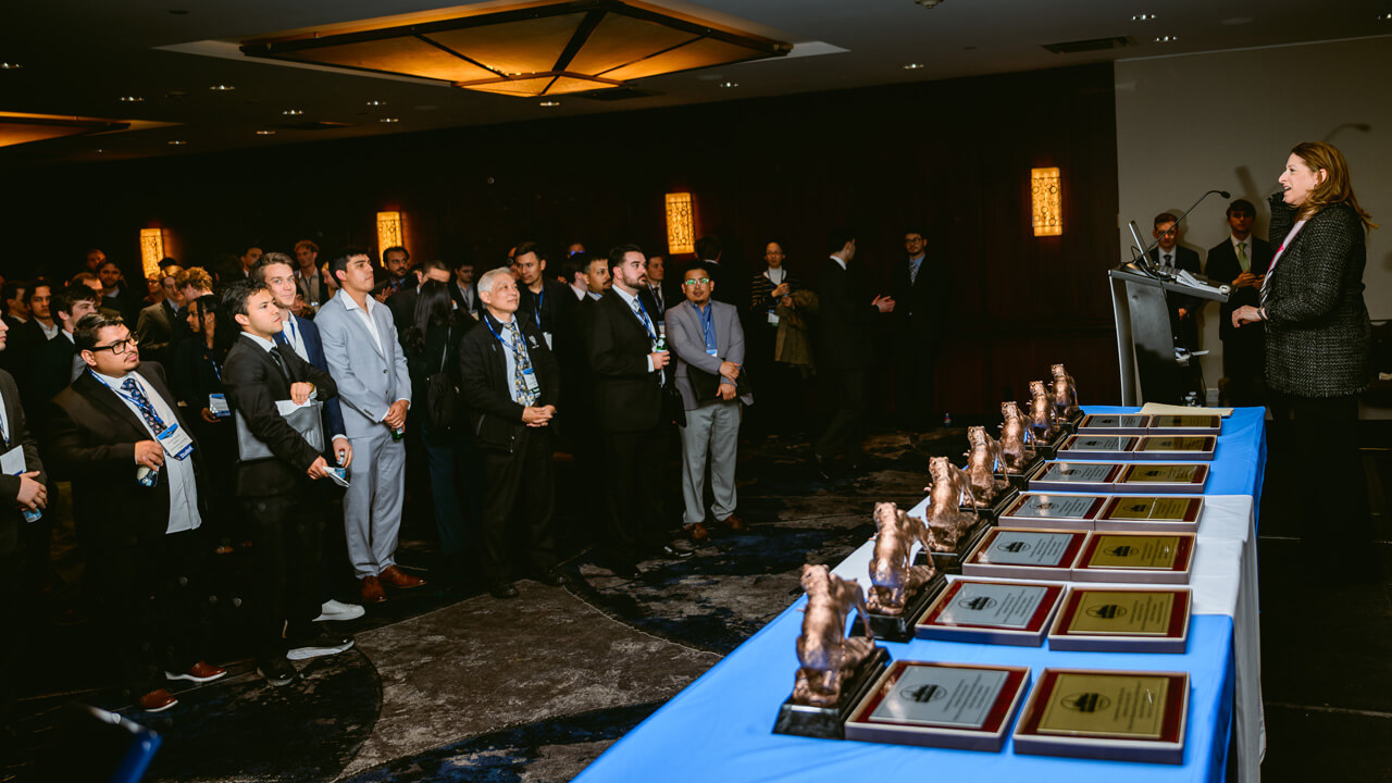 Dean Holly Raider speaks at a microphone next to a table with a blue tablecloth holding the trophies for the global portfolio competition winners.