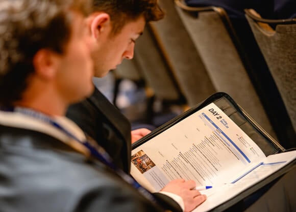 Two male students sitting in the Forum audience holding a program open to the Day 2 page.