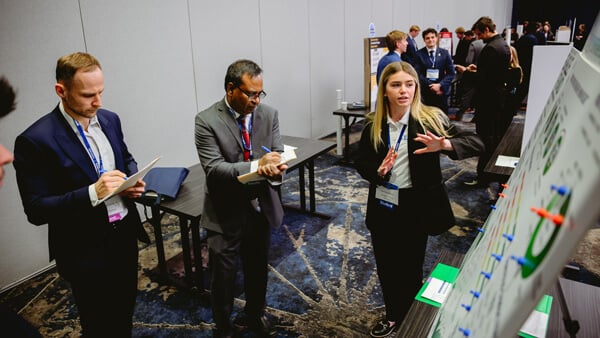 A student explains her poster during the Forum poster session, while two faculty members take notes on clipboards.