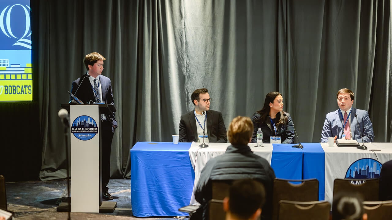 Benny Amarone standing at a podium next to a panel table where Dennis Kilic, Caroline King, and Parker Moorman are seated.