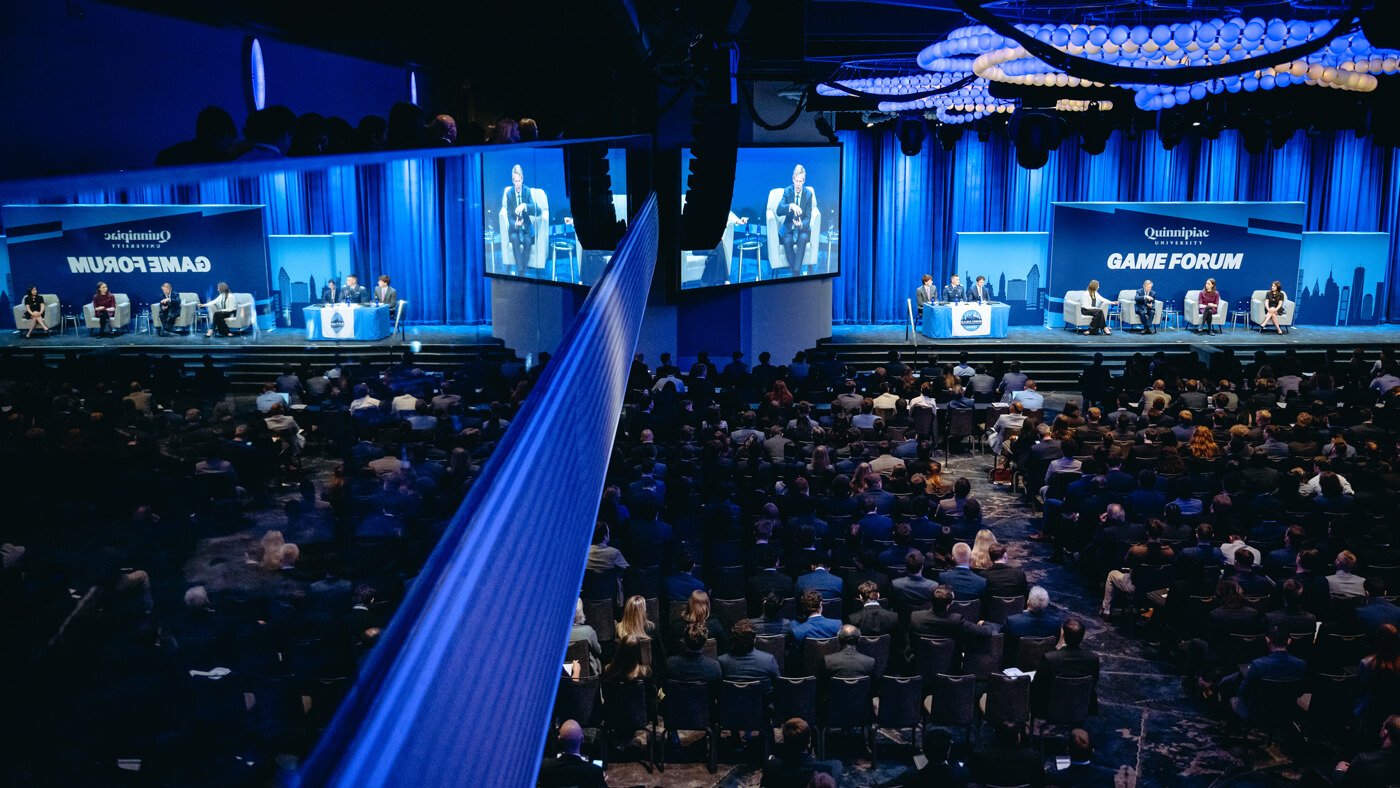 A mirrored view of the GAME Forum stage shows panelists engaged in conversation, with their reflections visible on the walls and ceiling.