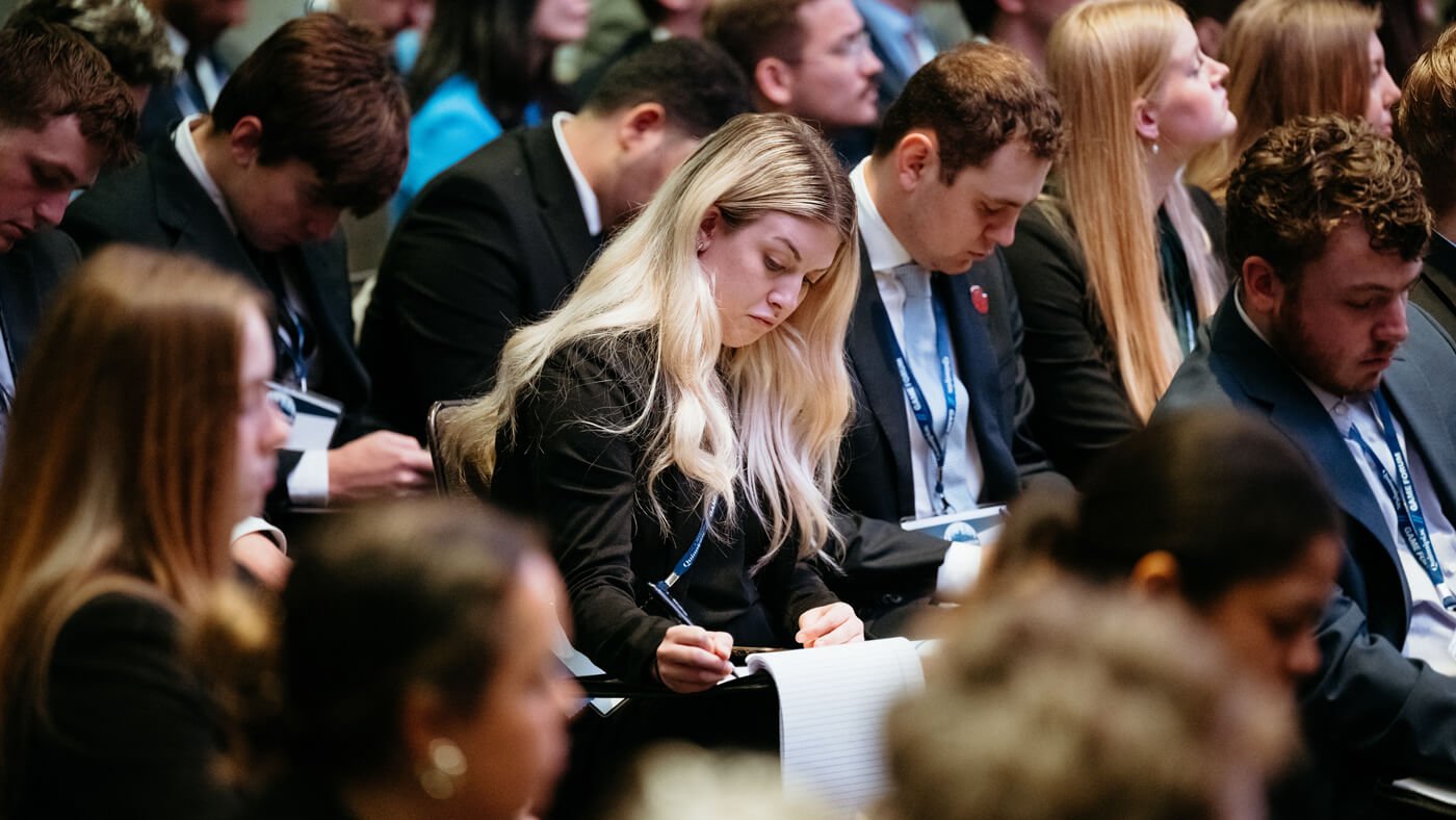 A focused student takes notes during a session, surrounded by rows of fellow attendees in business attire.