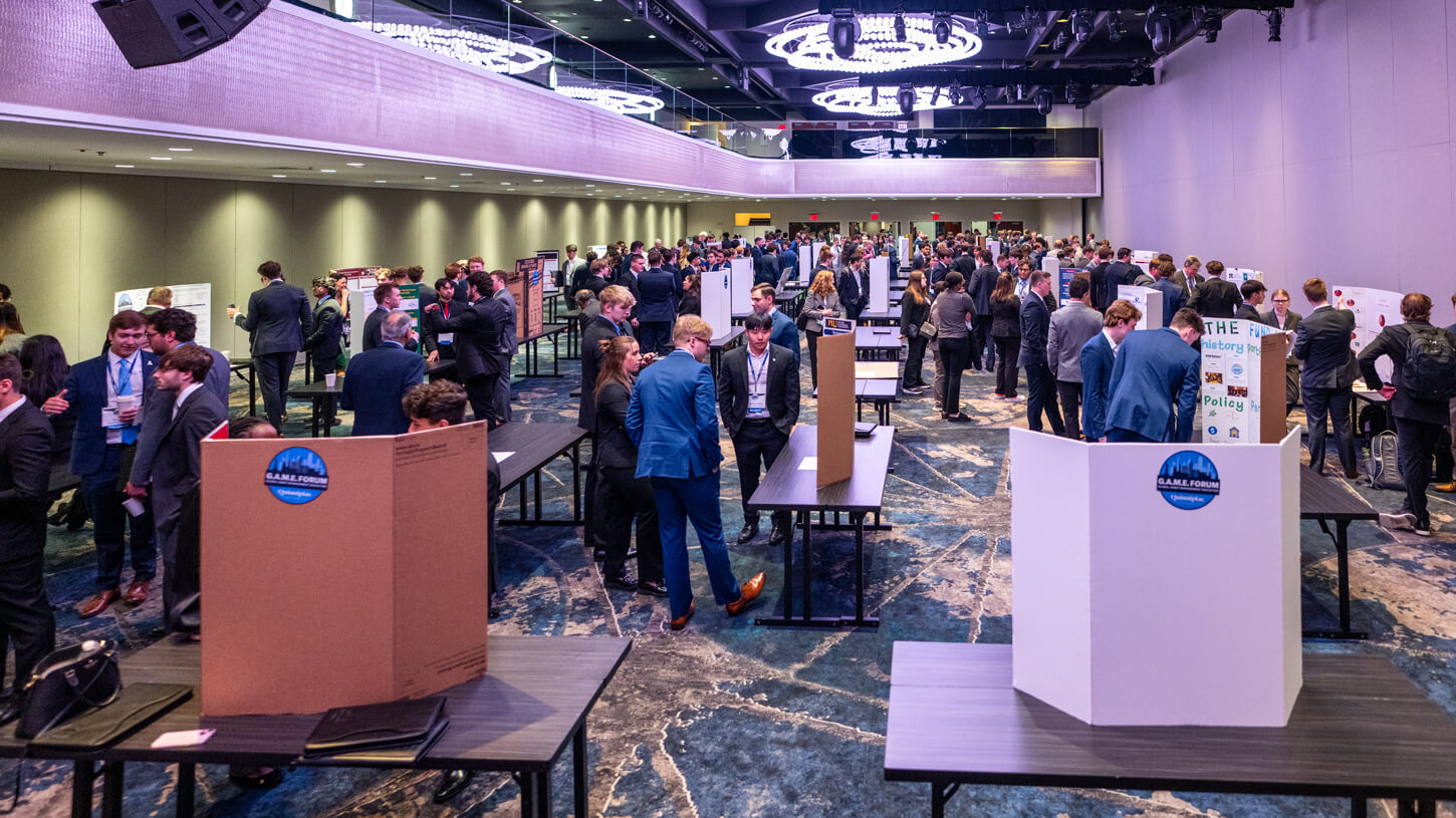 A wide view of the GAME Forum venue shows dozens of teams presenting posters at black tables, with a large crowd of students and judges filling the room.