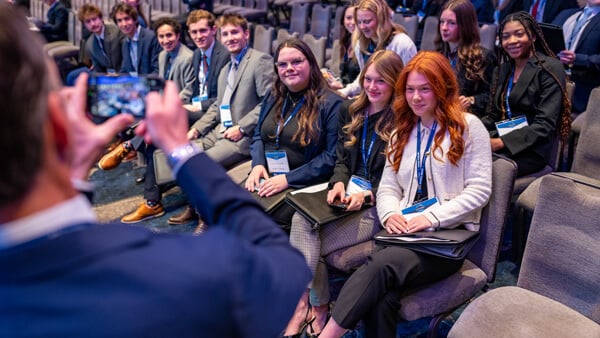 A row of eight students and another row of four students behind them sitting in the ballroom, smiling as a professor takes a photo.