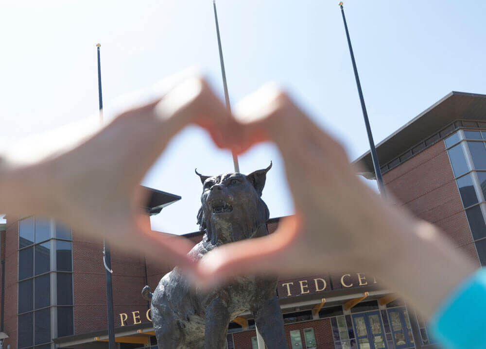 A student forms her hand into a heart with the bobcat statue in the background